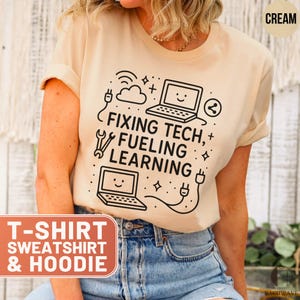 May include: Cream-colored t-shirt with the text "FIXING TECH, FUELING LEARNING" in black. The design includes laptop graphics, a wrench, and a plug. The shirt is part of a collection that includes sweatshirts and hoodies.