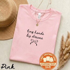 May include: A light pink t-shirt with the text "tiny hands big dreams" in black script and a bow graphic. A gold necklace with a pendant is on the shirt. A straw hat and dried plants are in the background.