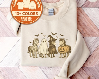 May include: Sand-colored sweatshirt featuring a Halloween-themed graphic of four cartoon horses dressed as a zebra, ghost, witch, and jack-o'-lantern. The sweatshirt has a crew neck and long sleeves. The image also includes text that says "10+ COLORS" and "FAST SHIPPING".