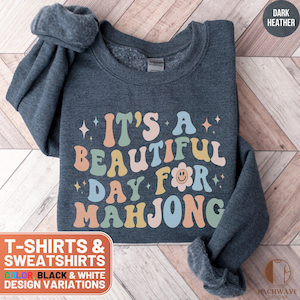 May include: Dark heather grey sweatshirt with the text "It's a beautiful day for Mahjong" in colorful retro font. The text is surrounded by small stars and a smiley face flower.