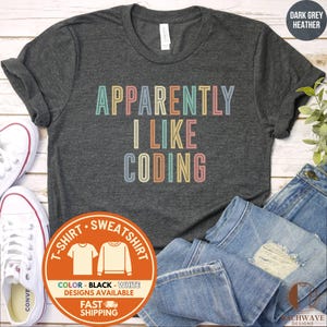 May include: Dark grey heather t-shirt with the text "APPARENTLY I LIKE CODING" in pastel colors. The image includes white sneakers, blue jeans, and a circular graphic with shirt and sweatshirt icons.
