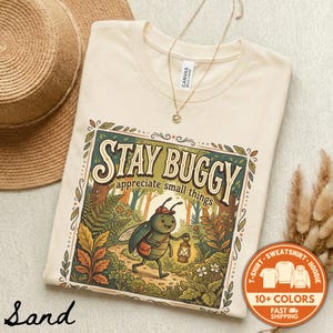May include: Sand-colored t-shirt with the words "STAY BUGGY appreciate small things" in a decorative frame. The graphic features a cartoon bug with a lantern walking through a forest. A straw hat and necklace are also visible.