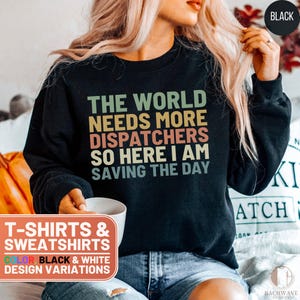 May include: A black sweatshirt with the text "The world needs more dispatchers so here I am saving the day" in a vintage style font. The text is in different colors, including green, orange, and blue. The sweatshirt is available in black and white.