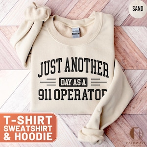 May include: Sand-colored sweatshirt with the text "JUST ANOTHER DAY AS A 911 OPERATOR" in black. The sweatshirt is folded, with the sleeves rolled up. The words "T-SHIRT SWEATSHIRT & HOODIE" are in a red box.
