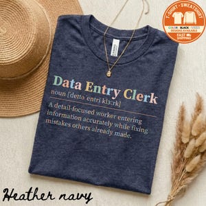 May include: A heather navy t-shirt with the text "Data Entry Clerk" in colorful letters. The definition of the job title is printed below the title. A gold necklace and a straw hat are next to the shirt.