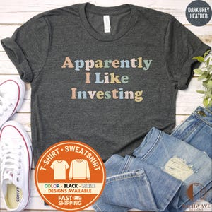 May include: Dark grey heather t-shirt with the text "Apparently I Like Investing" in pastel colors. The shirt is displayed with white sneakers and blue jeans. A circular graphic indicates t-shirt and sweatshirt options.