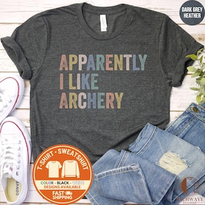 May include: A dark grey heather t-shirt featuring the text "APPARENTLY I LIKE ARCHERY" in a distressed, multi-colored font. The shirt is displayed alongside white canvas sneakers and blue denim jeans, with a circular graphic label in the corner.
