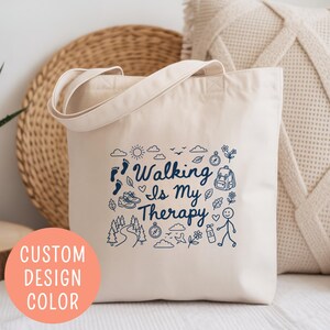 May include: Cream-colored tote bag with the text "Walking Is My Therapy" and various outdoor-themed illustrations in blue. The bag has a simple design with a handle and is made of a canvas-like material.