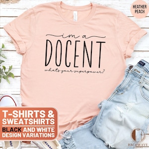 May include: A peach colored t-shirt with black text that reads "I'm a DOCENT whats your superpower?"  The t-shirt is on a wooden surface with a pair of blue jeans and a pair of white sneakers.