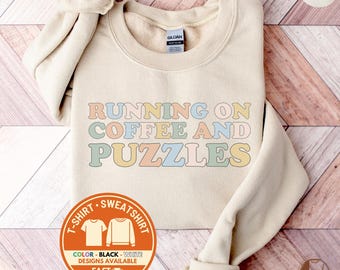 May include: Sand-colored sweatshirt with the text "RUNNING ON COFFEE AND PUZZLES" in pastel colors. The sweatshirt has a crew neck and long sleeves. The image also includes a graphic indicating t-shirt and sweatshirt designs.