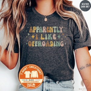 May include: Dark grey t-shirt with the text "APPARENTLY I LIKE OFFROADING" in retro-style lettering. The shirt has a crew neck and short sleeves. The design includes stars and a color palette of brown, yellow, green, and pink.
