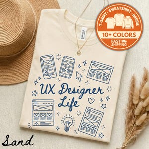 May include: Sand-colored t-shirt with a blue graphic design featuring smartphones, website layouts, and the text "UX Designer Life." The shirt includes a round orange graphic with the words "10+ COLORS" and "FAST SHIPPING."