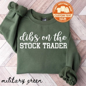 May include: Military green sweatshirt with the text "dibs on the STOCK TRADER" in white. The sweatshirt has a crew neck and long sleeves. A circular orange graphic with the words "CUSTOM TEXT" is in the upper right corner.