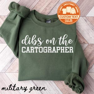 May include: Military green sweatshirt with the text "dibs on the CARTOGRAPHER" in white. The sweatshirt has a crew neck and long sleeves. A circular orange graphic with the words "Custom Text" is in the upper right corner.