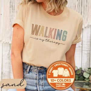 May include: A sand-colored t-shirt with the text "WALKING is my therapy" in a rainbow gradient. The shirt has rolled-up sleeves and is paired with blue jeans. A circular graphic indicates the shirt is available in 10+ colors.
