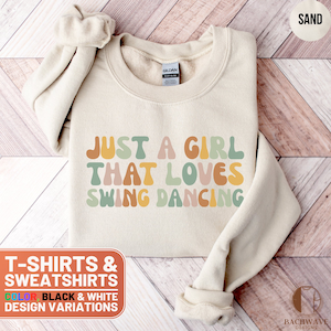 May include: A beige sweatshirt with the text "Just a girl that loves swing dancing" in colorful, retro-style lettering. The sweatshirt is available in black and white design variations.