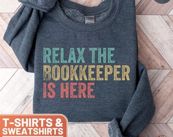 Puede incluir: Sudadera gris oscuro jaspeada con el texto "Relax the Bookkeeper is Here" en una fuente de estilo vintage con letras de colores. El texto está centrado en la sudadera.