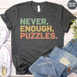 May include: A dark grey heather t-shirt with the text "NEVER. ENOUGH. PUZZLES." printed in a retro style with green, yellow, and orange colors.