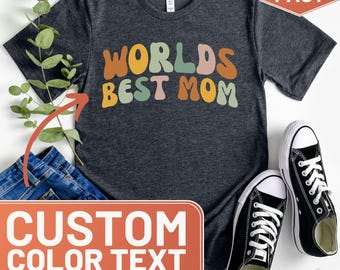 Puede incluir: Una camiseta gris oscuro con el texto "WORLDS BEST MOM" en una fuente ondulada de colores. El texto está en tonos de amarillo, naranja, verde, rosa y marrón. La camiseta se muestra sobre un fondo blanco con un par de zapatillas negras y un par de pantalones vaqueros azules.