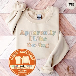 May include: A sand-colored crewneck sweatshirt featuring the text "Apparently I Like Coding" printed in a multi-colored, retro-style font. The garment has long sleeves, a ribbed collar, and a Gildan brand label visible at the inner neckline.
