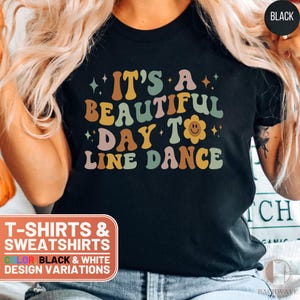 May include: Black t-shirt with the text "IT'S A BEAUTIFUL DAY TO LINE DANCE" in retro-style lettering. The design includes a smiling flower and star accents. The shirt is part of a collection of T-shirts and sweatshirts.