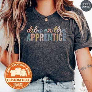 May include: Heather dark grey t-shirt with the text "dibs on the APPRENTICE" in colorful letters. The shirt has a crew neck and short sleeves. A logo with the words "CUSTOM TEXT" and "FAST SHIPPING" is visible.