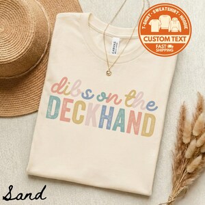 May include: A sand-colored t-shirt with the text "dibs on the DECKHAND" in pastel colors. The shirt has a crew neck and is made of a soft material. A gold necklace and a straw hat are also visible.