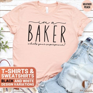May include: A pink t-shirt with the text "I'm a Baker, what's your superpower?" in black. The t-shirt is on a wooden surface with a pair of blue jeans and a pair of white sneakers.