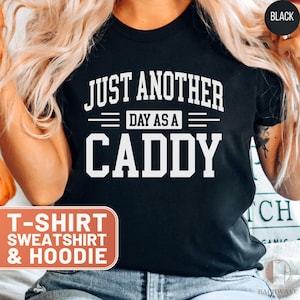 May include: Black t-shirt with the text "JUST ANOTHER DAY AS A CADDY" in white block letters. The shirt also has the words "T-SHIRT SWEATSHIRT & HOODIE" in a red and white box.