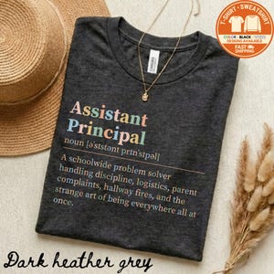 May include: Dark heather grey t-shirt with the words "Assistant Principal" in colorful text. The definition of Assistant Principal is printed below the title. A gold necklace and a straw hat are visible.