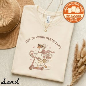 May include: A sand-colored t-shirt featuring a graphic of a goose wearing sunglasses and a scarf, riding a scooter while holding coffee cups and a bag. The text reads OFF TO WORK BESTIE DUTY. A gold-toned chain necklace rests on the collar.