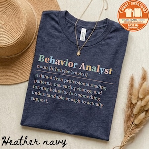 May include: Heather navy t-shirt with the text "Behavior Analyst" in colorful letters. The shirt includes a definition of the term. A gold necklace and a straw hat are also in the image.