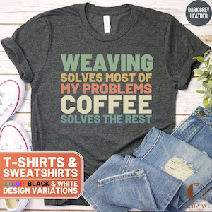 May include: A dark grey heather t-shirt with the text "WEAVING SOLVES MOST OF MY PROBLEMS COFFEE SOLVES THE REST" in a retro-style font with colorful letters. The text is centered on the shirt. The shirt is displayed on a wooden surface with a pair of jeans and a pair of white sneakers.