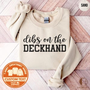 May include: Sand-colored sweatshirt with the text "dibs on the DECKHAND" in black. The sweatshirt has a crew neck and long sleeves. A circular graphic at the bottom left indicates the product is a t-shirt, sweatshirt, or hoodie. The word "SAND" is in the top right corner.