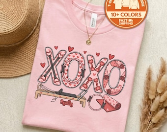 May include: Pink t-shirt with a graphic design featuring the letters "XOXO" filled with hearts and dumbbells. The design includes a Pilates reformer, a yoga mat, and a ring. The shirt has a round neck and short sleeves.