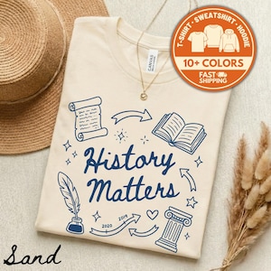 May include: A sand-colored t-shirt with the words "History Matters" in navy blue, surrounded by historical illustrations. The shirt features a round orange sticker with text and a straw hat in the background.