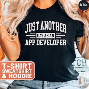 May include: Black t-shirt with white text that reads "JUST ANOTHER DAY AS AN APP DEVELOPER." The shirt also has the words "T-SHIRT SWEATSHIRT & HOODIE" in a separate box. The shirt is a crew neck.
