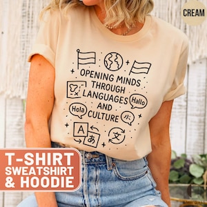 May include: Cream-colored t-shirt with the text "OPENING MINDS THROUGH LANGUAGES AND CULTURE" in black. The design includes flags, a globe, speech bubbles with "Hola" and "Hallo", and other language symbols. The shirt is part of a T-shirt, sweatshirt, and hoodie collection.