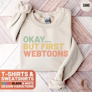 May include: A sand-colored sweatshirt with the text "OKAY... BUT FIRST WEBTOONS" in a rainbow gradient. The text is in a retro font style. The sweatshirt is available in black and white design variations.