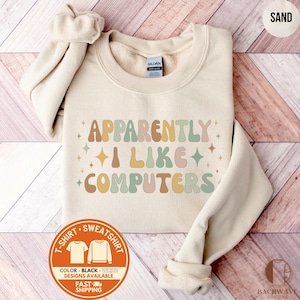 May include: A sand-colored sweatshirt with the text "APPARENTLY I LIKE COMPUTERS" in retro-style lettering. The design includes colorful text and star accents. The sweatshirt is a Gildan brand and features a crew neck.