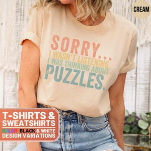 May include: A cream colored t-shirt with the text "SORRY... I WASN'T LISTENING. I WAS THINKING ABOUT PUZZLES." printed in a vintage style font. The text is in shades of pink, yellow, and blue. The t-shirt is available in black and white.