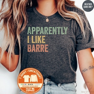 May include: Heather dark grey t-shirt with the text "Apparently I Like Barre" in green, yellow, and orange. The shirt has a crew neck and short sleeves. A circular logo at the bottom indicates t-shirt and sweatshirt designs are available.