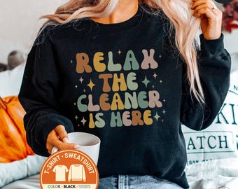 May include: Black sweatshirt with the text "RELAX THE CLEANER IS HERE" in retro-style lettering. The design includes stars and a color palette of brown, green, pink, and yellow. A white mug is held in the hand. The shirt is available in black and white.