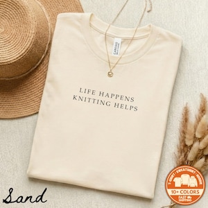 May include: A sand-colored t-shirt featuring the black text "LIFE HAPPENS KNITTING HELPS" printed on the chest. The shirt has a crew neckline and a small tag visible at the inner collar. A delicate gold-toned chain necklace rests on the fabric.