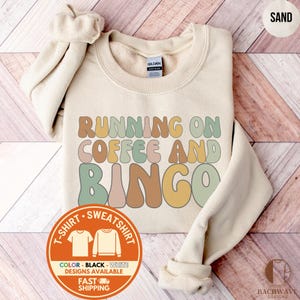 May include: Sand-colored sweatshirt with the text "RUNNING ON COFFEE AND BINGO" in retro-style lettering. The design includes a round graphic with color options and fast shipping information. A Gildan tag is visible.