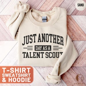 May include: Sand-colored sweatshirt with the text "JUST ANOTHER DAY AS A TALENT SCOUT" in black. The sweatshirt is a Gildan brand and has a crew neck. The image also includes a graphic that says "T-SHIRT SWEATSHIRT & HOODIE".