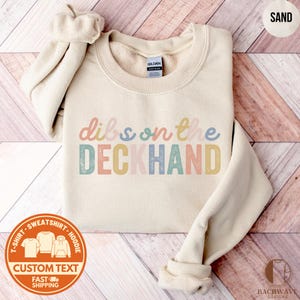 May include: Sand-colored sweatshirt with the text "dibs on the DECKHAND" in pastel colors. The sweatshirt has a crew neck and long sleeves. A circular graphic at the bottom reads "T-SHIRT - SWEATSHIRT - HOODIE" and "CUSTOM TEXT" with a fast shipping icon.