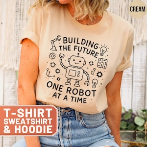 May include: Cream-colored t-shirt with the text "BUILDING THE FUTURE ONE ROBOT AT A TIME" and a graphic of a robot. The shirt also has the words "T-SHIRT SWEATSHIRT & HOODIE" in a rectangular box.