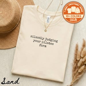May include: A sand-colored t-shirt with the text "silently judging your pilates form" in black. A gold necklace and a straw hat are visible. A circular orange sticker indicates multiple colors and fast shipping.