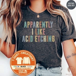 May include: A heather dark grey t-shirt featuring the text "APPARENTLY I LIKE ACID ETCHING" in a multi-colored, retro-inspired font. The shirt has short sleeves and a crew neckline. A circular orange graphic overlay displays clothing options.